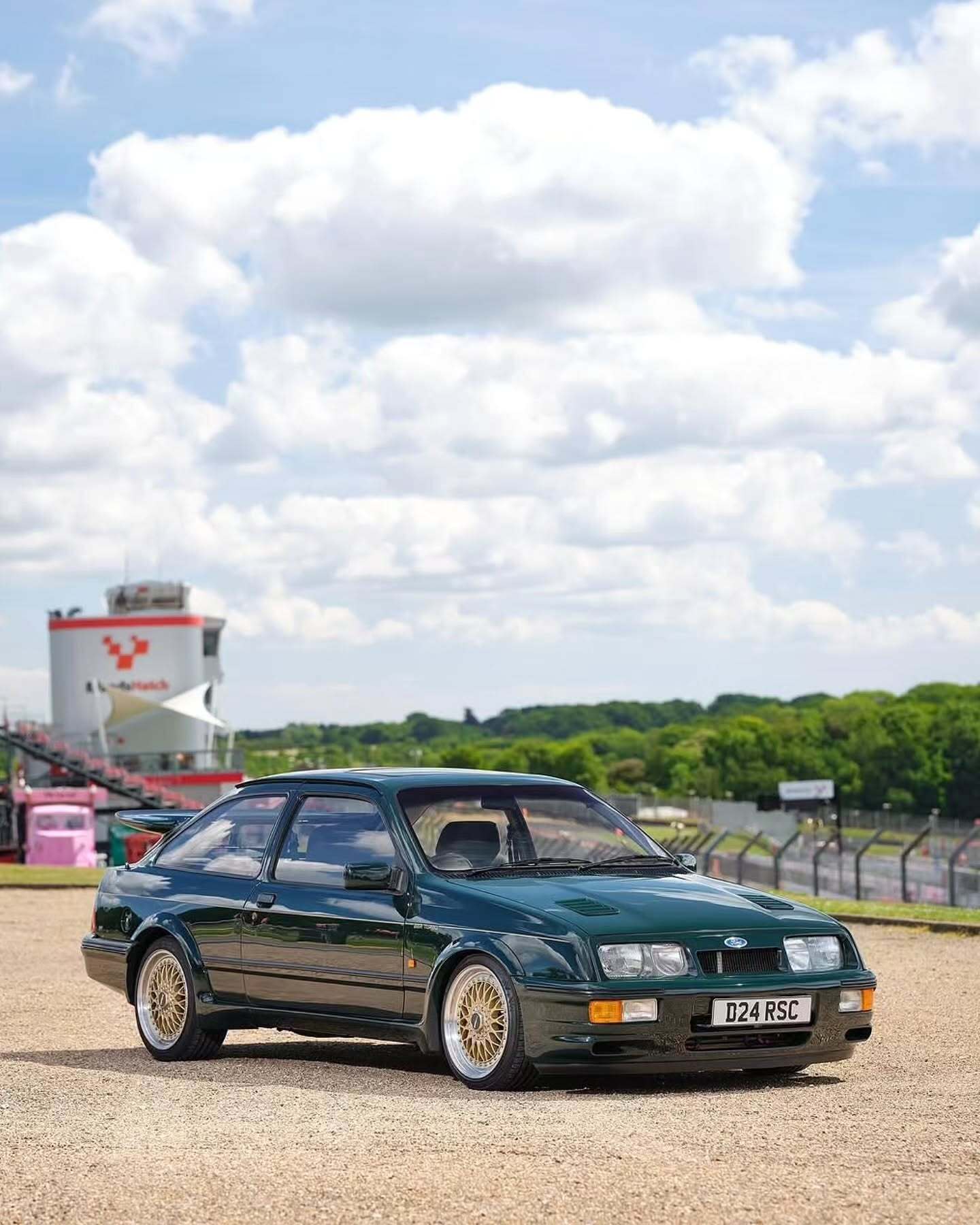 تصویر تمام‌قد و با زاویه باز از خودروی کلاسیک و اسپرت فورد سیرا کاسورث (Ford Sierra Cosworth) با رنگ تیره، که باله عظیم عقب و گلگیرهای برجسته آن در دهه ۱۹۸۰ میلادی آن را به یک نماد سرعت و سلطه بر پیست‌های مسابقه تبدیل کرد.
