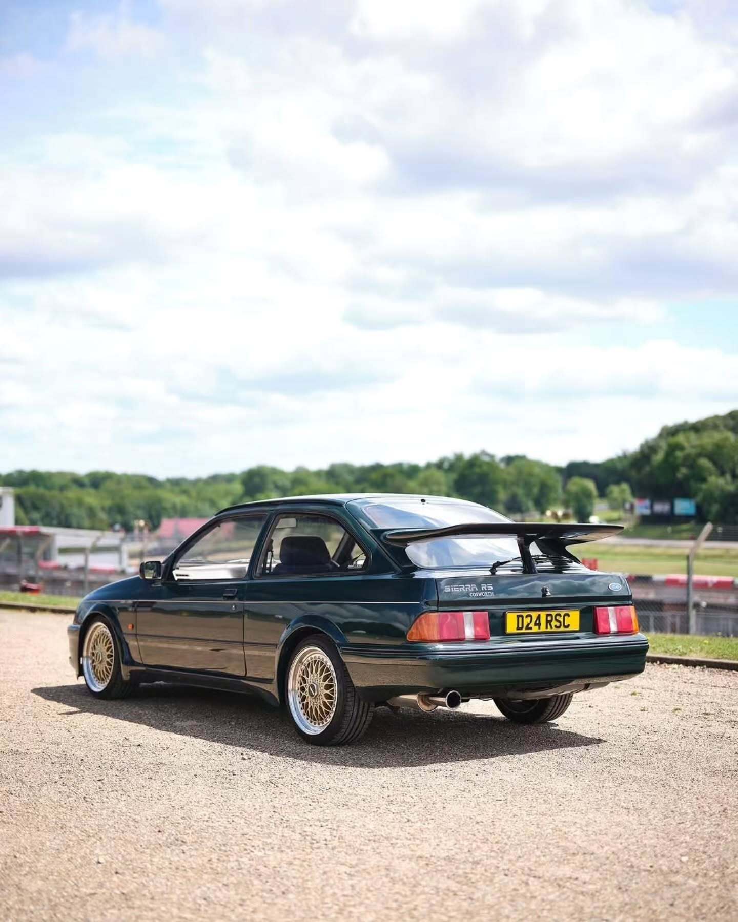 تصویر تمام‌قد و با زاویه باز از خودروی کلاسیک و اسپرت فورد سیرا کاسورث (Ford Sierra Cosworth) با رنگ تیره، که باله عظیم عقب و گلگیرهای برجسته آن در دهه ۱۹۸۰ میلادی آن را به یک نماد سرعت و سلطه بر پیست‌های مسابقه تبدیل کرد.