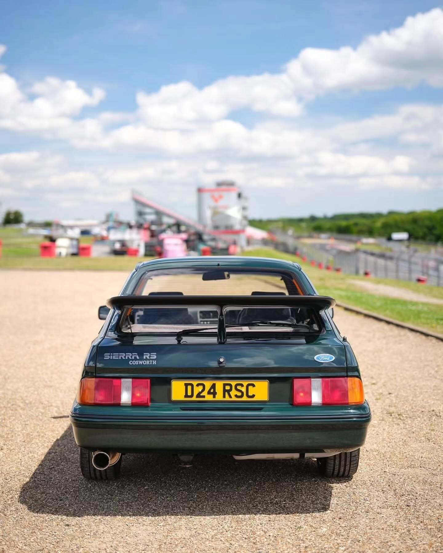 تصویر تمام‌قد و با زاویه باز از خودروی کلاسیک و اسپرت فورد سیرا کاسورث (Ford Sierra Cosworth) با رنگ تیره، که باله عظیم عقب و گلگیرهای برجسته آن در دهه ۱۹۸۰ میلادی آن را به یک نماد سرعت و سلطه بر پیست‌های مسابقه تبدیل کرد.