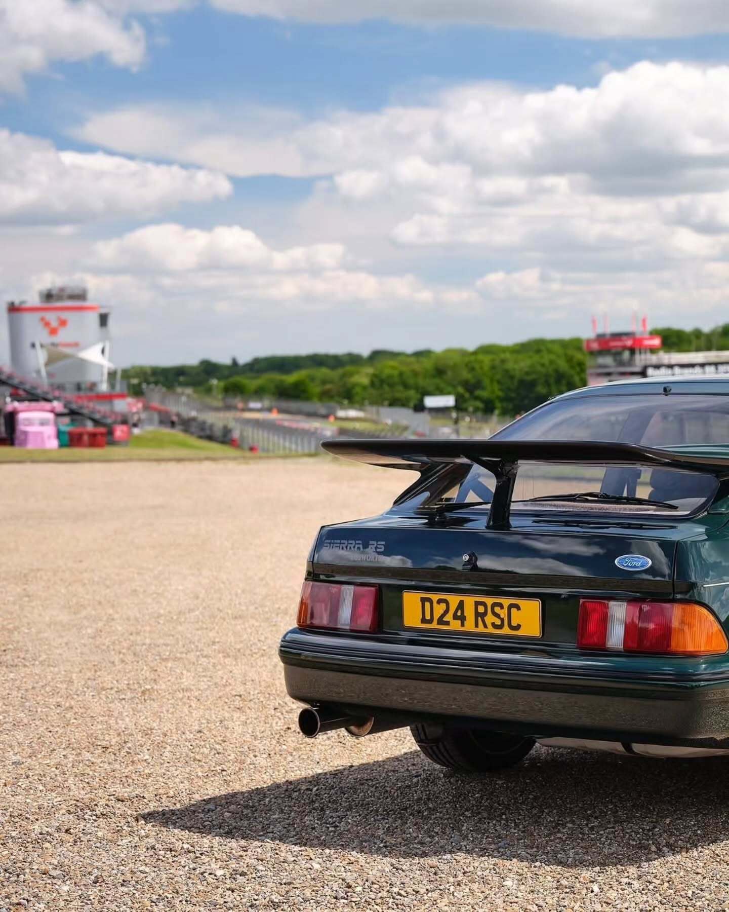 تصویر تمام‌قد و با زاویه باز از خودروی کلاسیک و اسپرت فورد سیرا کاسورث (Ford Sierra Cosworth) با رنگ تیره، که باله عظیم عقب و گلگیرهای برجسته آن در دهه ۱۹۸۰ میلادی آن را به یک نماد سرعت و سلطه بر پیست‌های مسابقه تبدیل کرد.