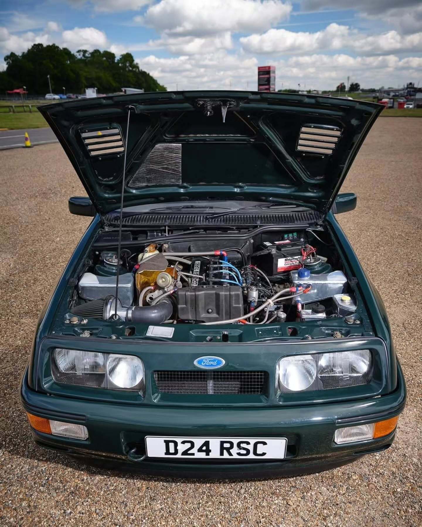 تصویر تمام‌قد و با زاویه باز از خودروی کلاسیک و اسپرت فورد سیرا کاسورث (Ford Sierra Cosworth) با رنگ تیره، که باله عظیم عقب و گلگیرهای برجسته آن در دهه ۱۹۸۰ میلادی آن را به یک نماد سرعت و سلطه بر پیست‌های مسابقه تبدیل کرد.