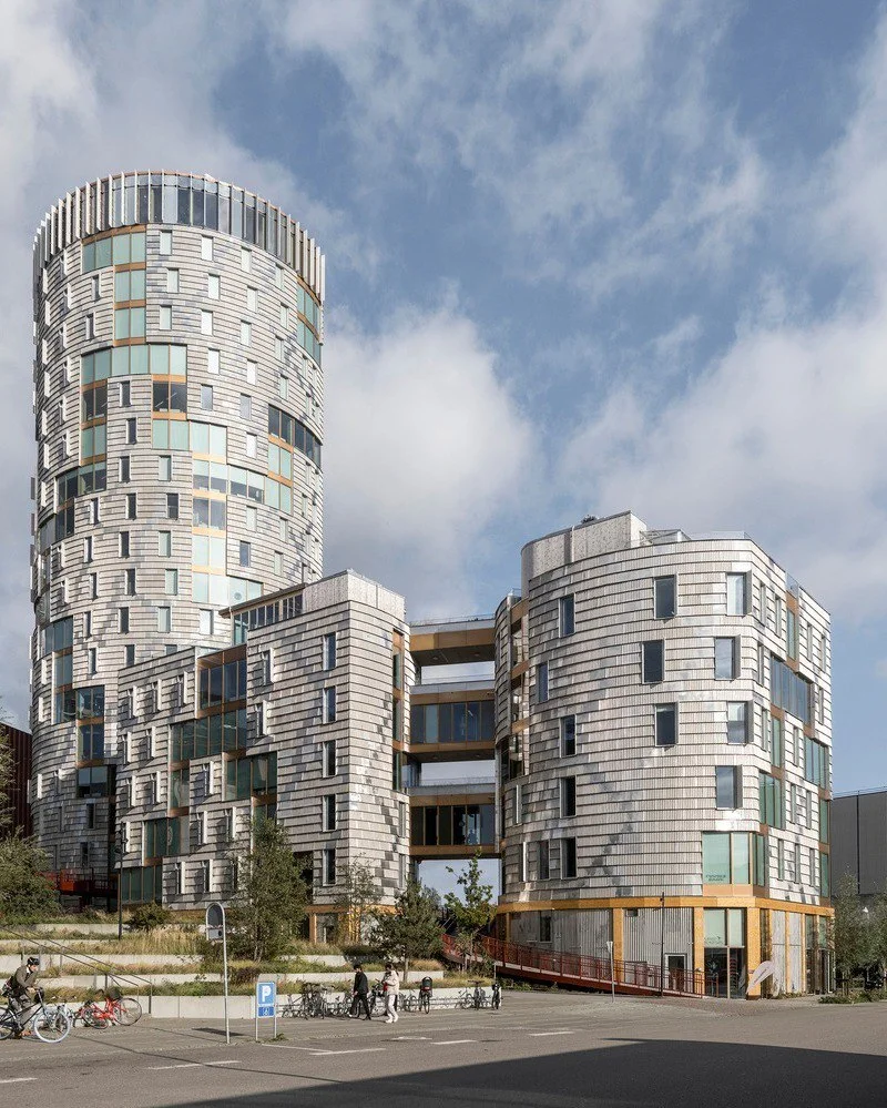 تصویر نمای خارجی و مدرن ساختمان TRÆ (ترا) در بندر آرهوس، دانمارک، که با سیستم سازهای چوبی-هیبریدی و پوشش نمای آلومینیوم بازیافتی توسط Lendager Arkitekter به سبک معماری چرخهمحور طراحی شده است. تصویر نمای خارجی و مدرن ساختمان TRÆ (ترا) در بندر آرهوس، دانمارک، که با سیستم سازهای چوبی-هیبریدی و پوشش نمای آلومینیوم بازیافتی توسط Lendager Arkitekter به سبک معماری چرخهمحور طراحی شده است.