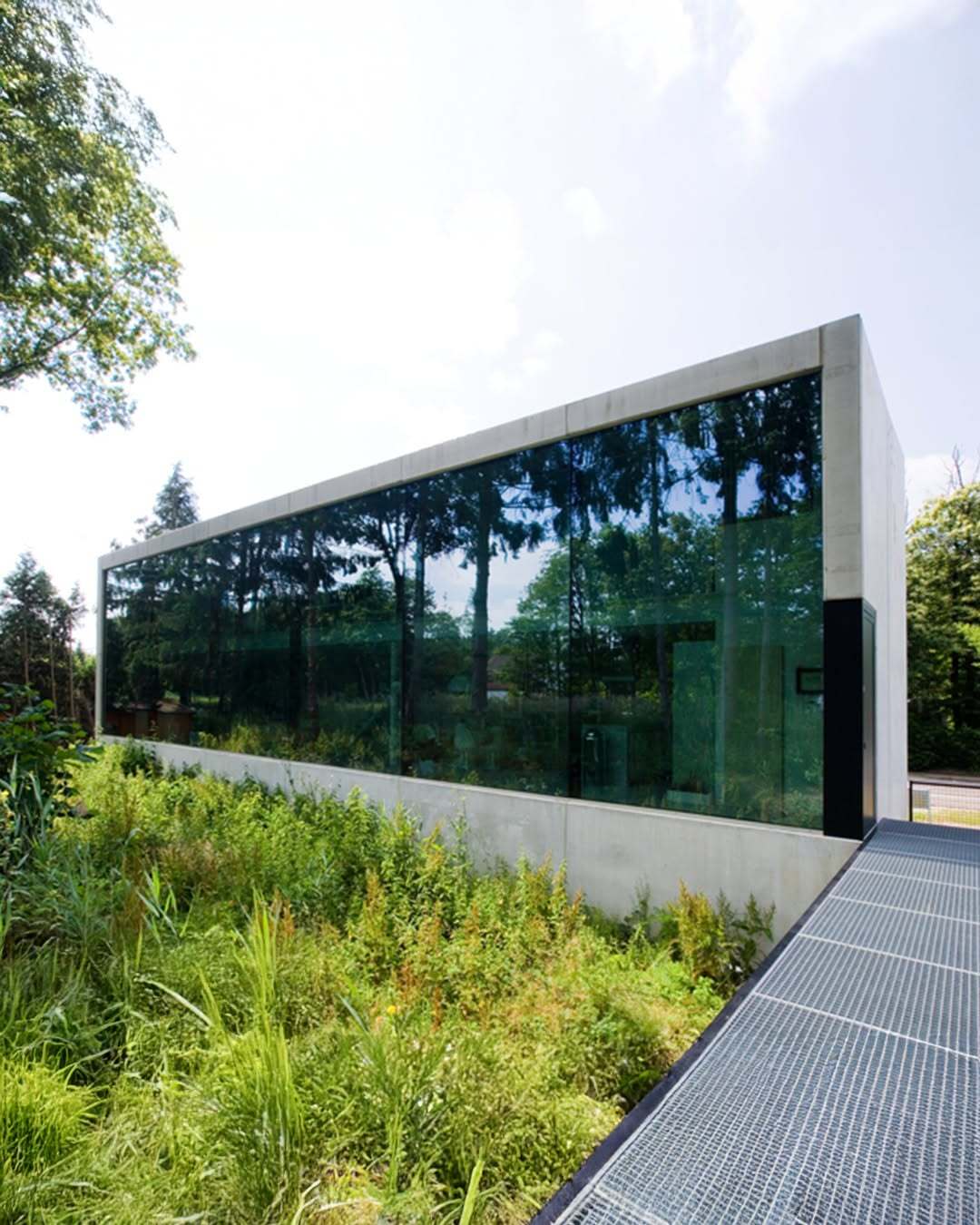 تصویر معماری مدرن و خطی یک خانه در بلژیک که نمای جلویی آن توسط بتن مستحکم پوشیده شده است، اما نمای پشتی کاملاً شیشه‌ای بوده و چشم‌اندازی بی‌نظیر به کانال آبی «کِمپیشه» را به نمایش می‌گذارد.