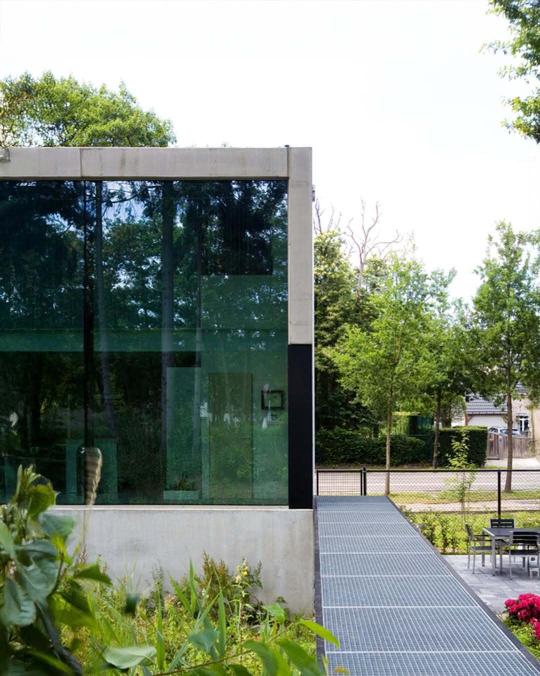 تصویر معماری مدرن و خطی یک خانه در بلژیک که نمای جلویی آن توسط بتن مستحکم پوشیده شده است، اما نمای پشتی کاملاً شیشه‌ای بوده و چشم‌اندازی بی‌نظیر به کانال آبی «کِمپیشه» را به نمایش می‌گذارد.