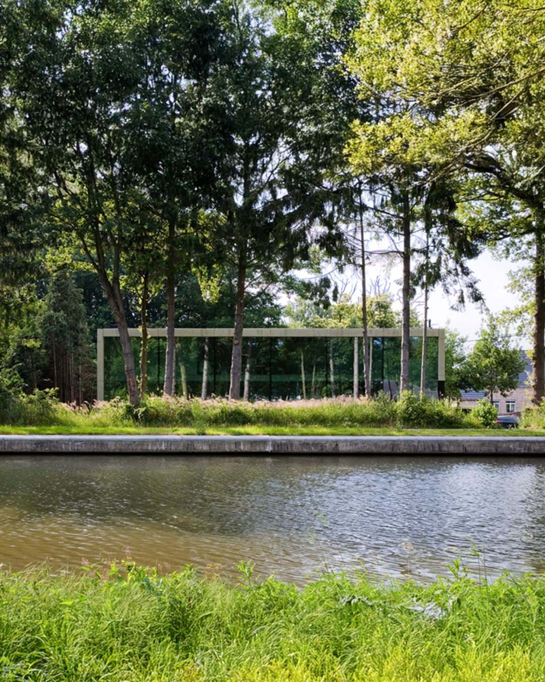 تصویر معماری مدرن و خطی یک خانه در بلژیک که نمای جلویی آن توسط بتن مستحکم پوشیده شده است، اما نمای پشتی کاملاً شیشه‌ای بوده و چشم‌اندازی بی‌نظیر به کانال آبی «کِمپیشه» را به نمایش می‌گذارد.