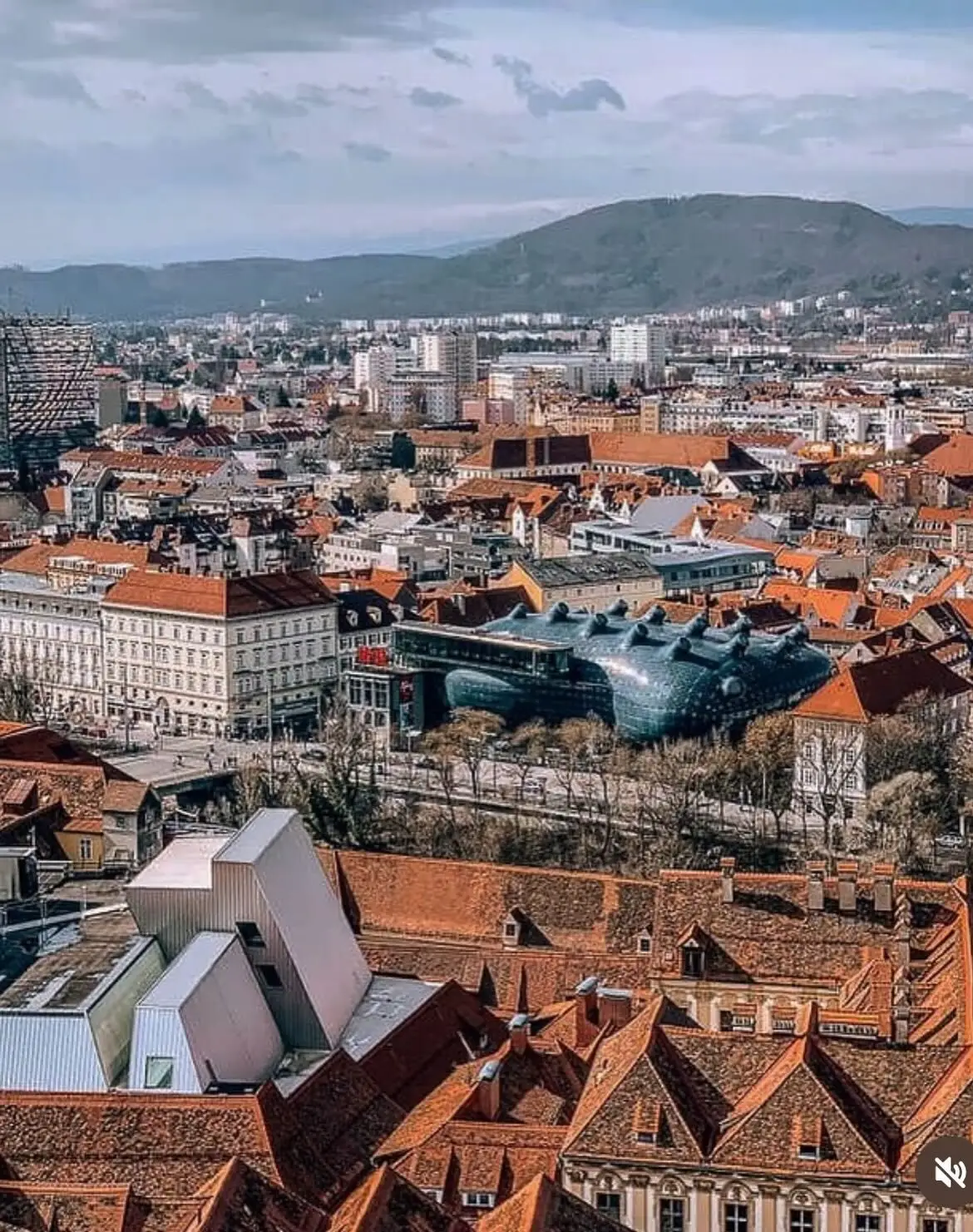 تصویری از نمای خارجی ساختمان موزه هنر کونست‌هاوس (Kunsthaus) در گراتس اتریش، که ساختار ارگانیک و غیرمعمول آن شبیه به یک موجود زنده با پوسته آبی رنگ است و به عنوان یک نماد معماری مدرن و «بیگانه دوست» طراحی شده توسط کولین فورنیر و پیتر کوک شناخته می‌شود.