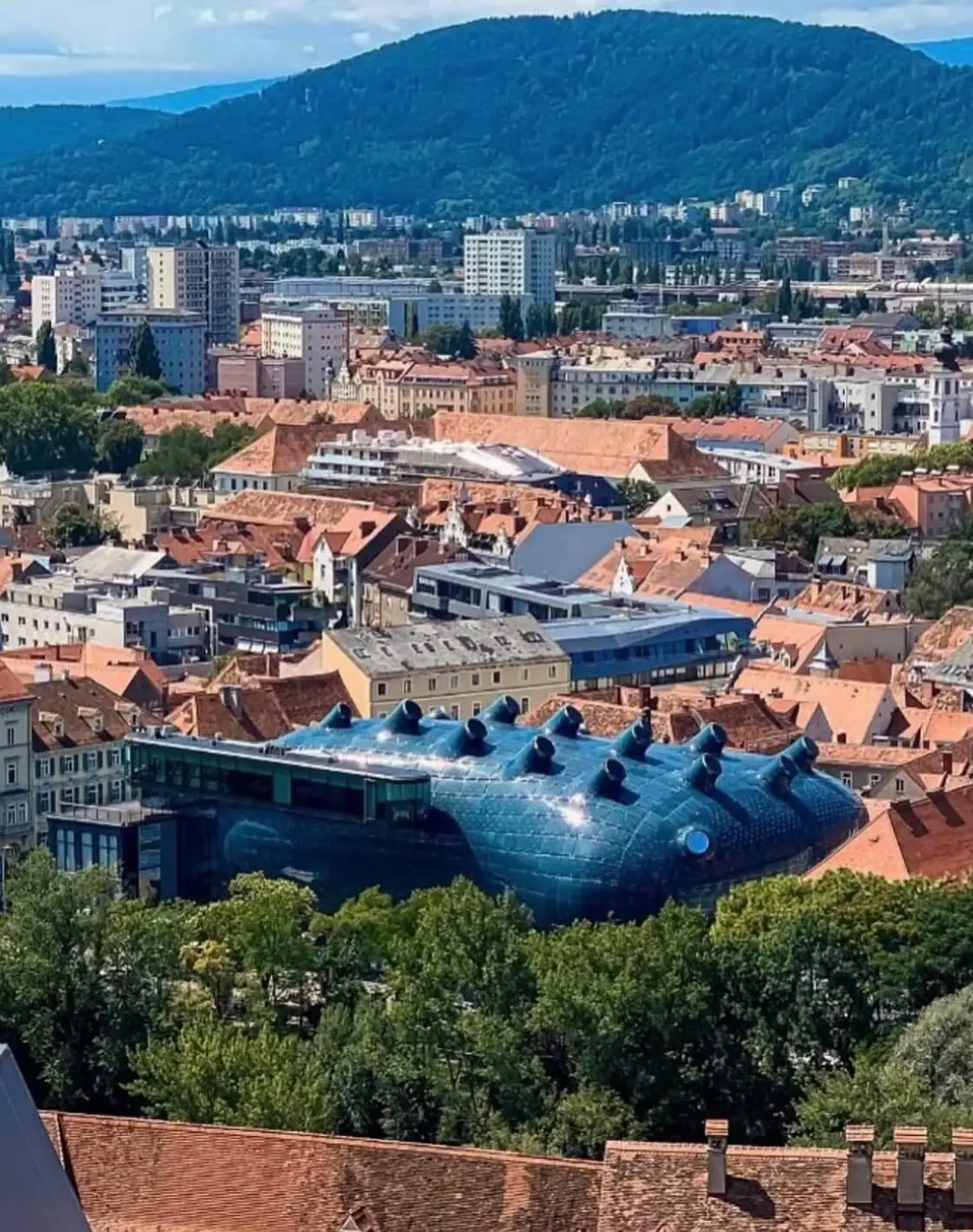 تصویری از نمای خارجی ساختمان موزه هنر کونست‌هاوس (Kunsthaus) در گراتس اتریش، که ساختار ارگانیک و غیرمعمول آن شبیه به یک موجود زنده با پوسته آبی رنگ است و به عنوان یک نماد معماری مدرن و «بیگانه دوست» طراحی شده توسط کولین فورنیر و پیتر کوک شناخته می‌شود.