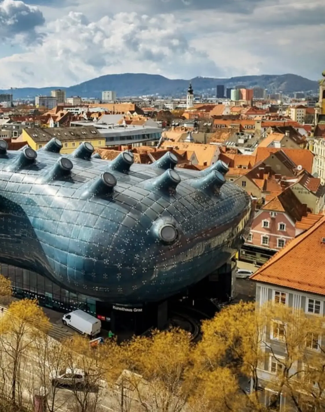 تصویری از نمای خارجی ساختمان موزه هنر کونست‌هاوس (Kunsthaus) در گراتس اتریش، که ساختار ارگانیک و غیرمعمول آن شبیه به یک موجود زنده با پوسته آبی رنگ است و به عنوان یک نماد معماری مدرن و «بیگانه دوست» طراحی شده توسط کولین فورنیر و پیتر کوک شناخته می‌شود.