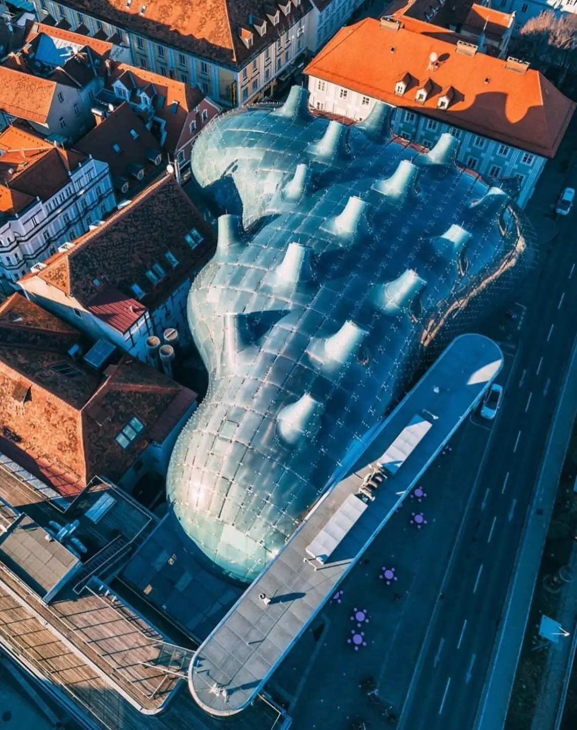 تصویری از نمای خارجی ساختمان موزه هنر کونست‌هاوس (Kunsthaus) در گراتس اتریش، که ساختار ارگانیک و غیرمعمول آن شبیه به یک موجود زنده با پوسته آبی رنگ است و به عنوان یک نماد معماری مدرن و «بیگانه دوست» طراحی شده توسط کولین فورنیر و پیتر کوک شناخته می‌شود.