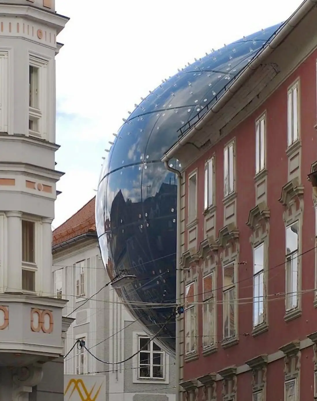 تصویری از نمای خارجی ساختمان موزه هنر کونست‌هاوس (Kunsthaus) در گراتس اتریش، که ساختار ارگانیک و غیرمعمول آن شبیه به یک موجود زنده با پوسته آبی رنگ است و به عنوان یک نماد معماری مدرن و «بیگانه دوست» طراحی شده توسط کولین فورنیر و پیتر کوک شناخته می‌شود.
