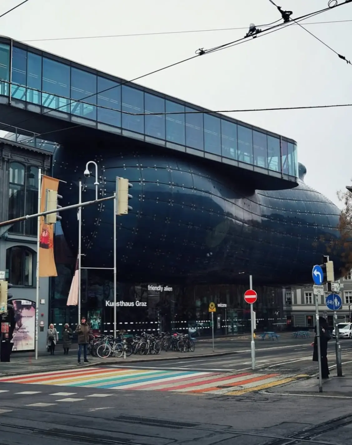 تصویری از نمای خارجی ساختمان موزه هنر کونست‌هاوس (Kunsthaus) در گراتس اتریش، که ساختار ارگانیک و غیرمعمول آن شبیه به یک موجود زنده با پوسته آبی رنگ است و به عنوان یک نماد معماری مدرن و «بیگانه دوست» طراحی شده توسط کولین فورنیر و پیتر کوک شناخته می‌شود.