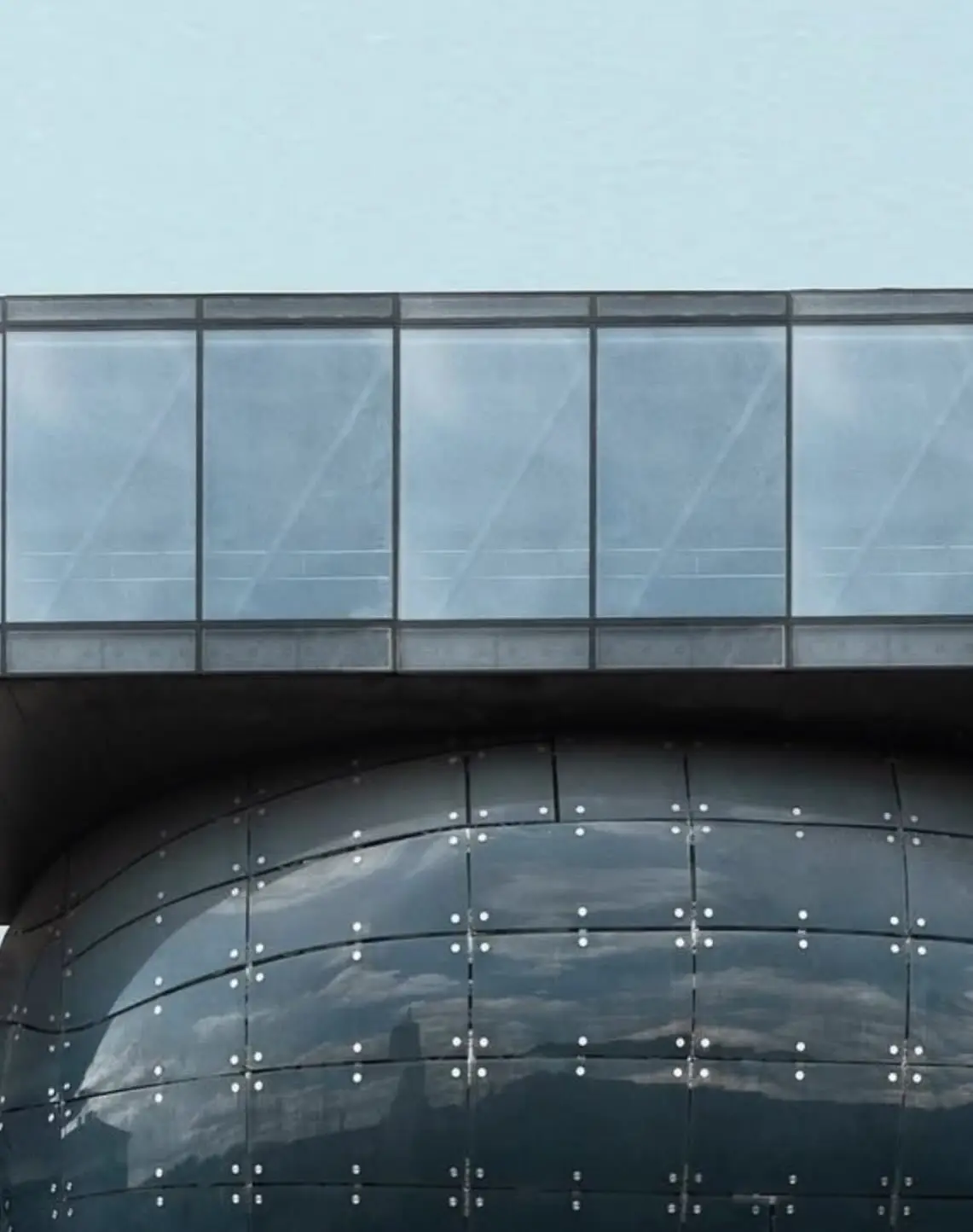 تصویری از نمای خارجی ساختمان موزه هنر کونست‌هاوس (Kunsthaus) در گراتس اتریش، که ساختار ارگانیک و غیرمعمول آن شبیه به یک موجود زنده با پوسته آبی رنگ است و به عنوان یک نماد معماری مدرن و «بیگانه دوست» طراحی شده توسط کولین فورنیر و پیتر کوک شناخته می‌شود.