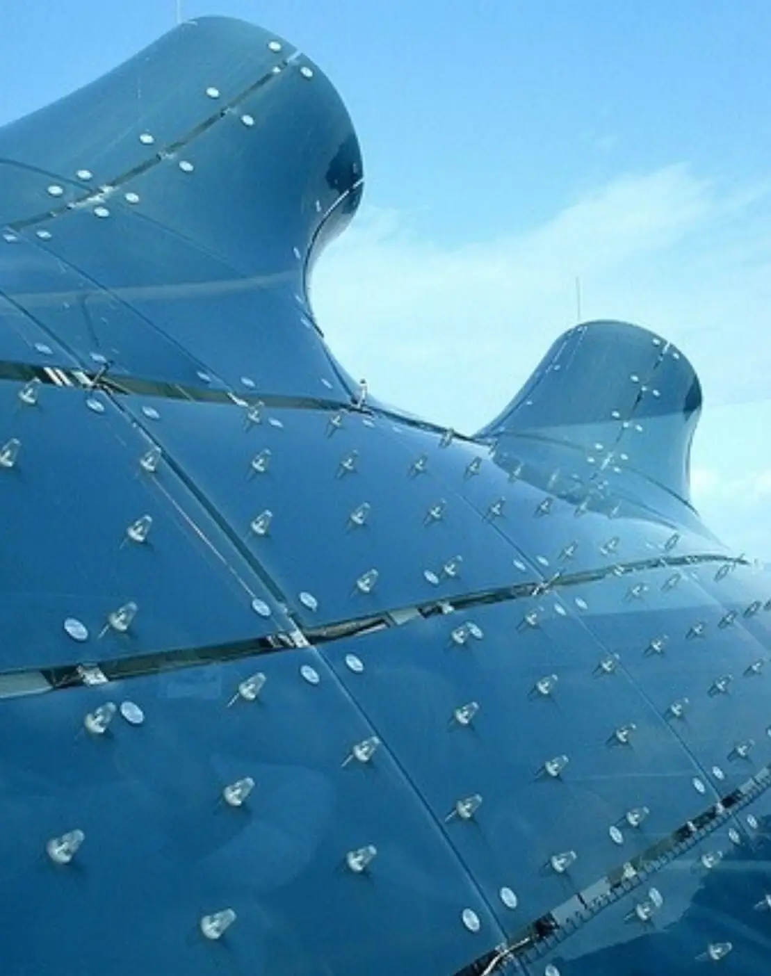 تصویری از نمای خارجی ساختمان موزه هنر کونست‌هاوس (Kunsthaus) در گراتس اتریش، که ساختار ارگانیک و غیرمعمول آن شبیه به یک موجود زنده با پوسته آبی رنگ است و به عنوان یک نماد معماری مدرن و «بیگانه دوست» طراحی شده توسط کولین فورنیر و پیتر کوک شناخته می‌شود.