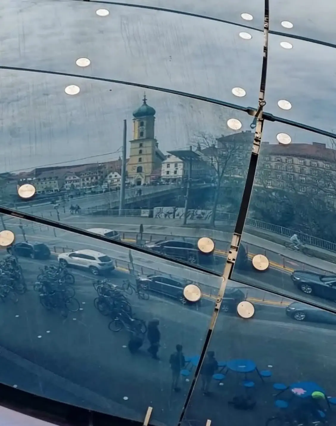 تصویری از نمای خارجی ساختمان موزه هنر کونست‌هاوس (Kunsthaus) در گراتس اتریش، که ساختار ارگانیک و غیرمعمول آن شبیه به یک موجود زنده با پوسته آبی رنگ است و به عنوان یک نماد معماری مدرن و «بیگانه دوست» طراحی شده توسط کولین فورنیر و پیتر کوک شناخته می‌شود.