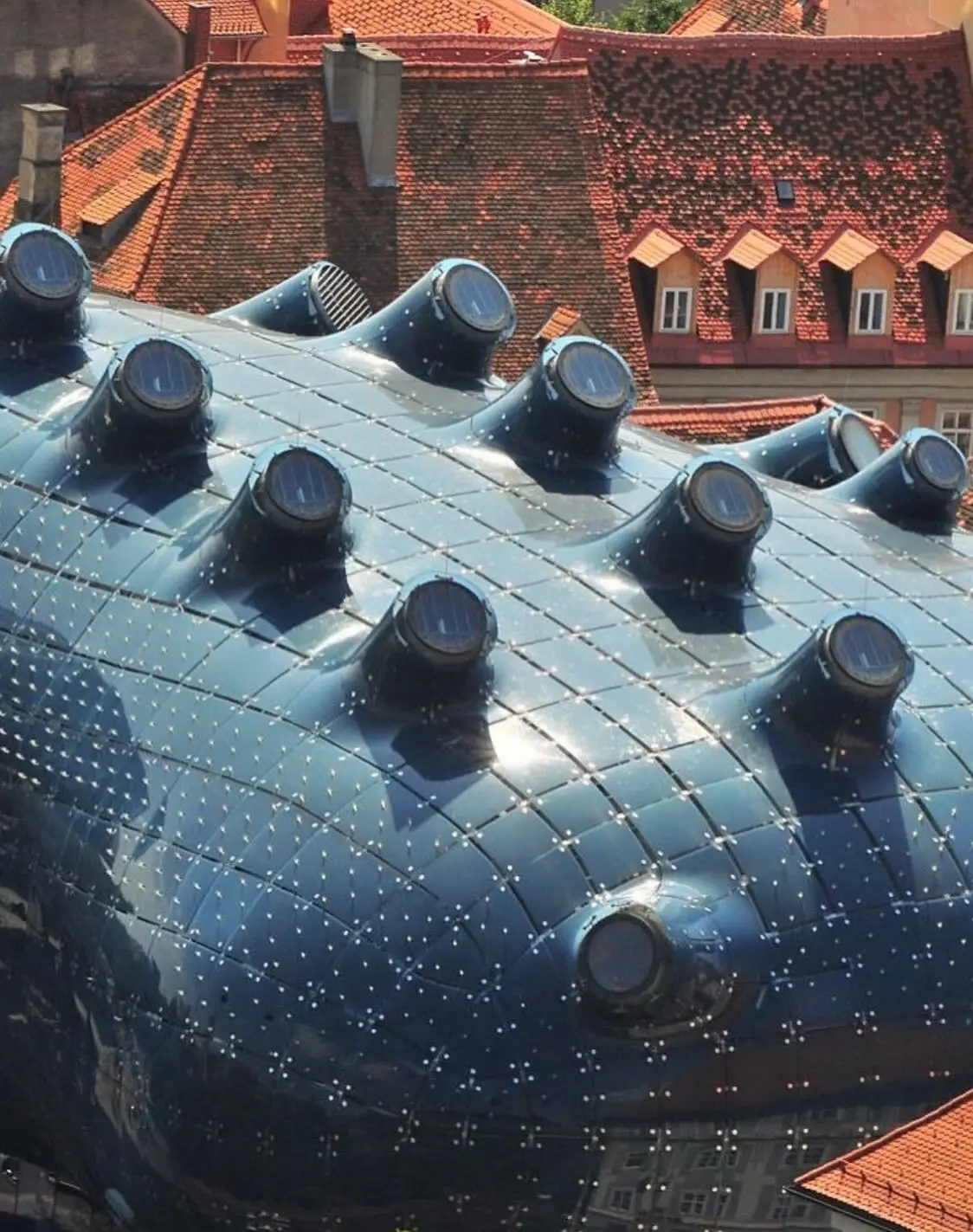 تصویری از نمای خارجی ساختمان موزه هنر کونست‌هاوس (Kunsthaus) در گراتس اتریش، که ساختار ارگانیک و غیرمعمول آن شبیه به یک موجود زنده با پوسته آبی رنگ است و به عنوان یک نماد معماری مدرن و «بیگانه دوست» طراحی شده توسط کولین فورنیر و پیتر کوک شناخته می‌شود.