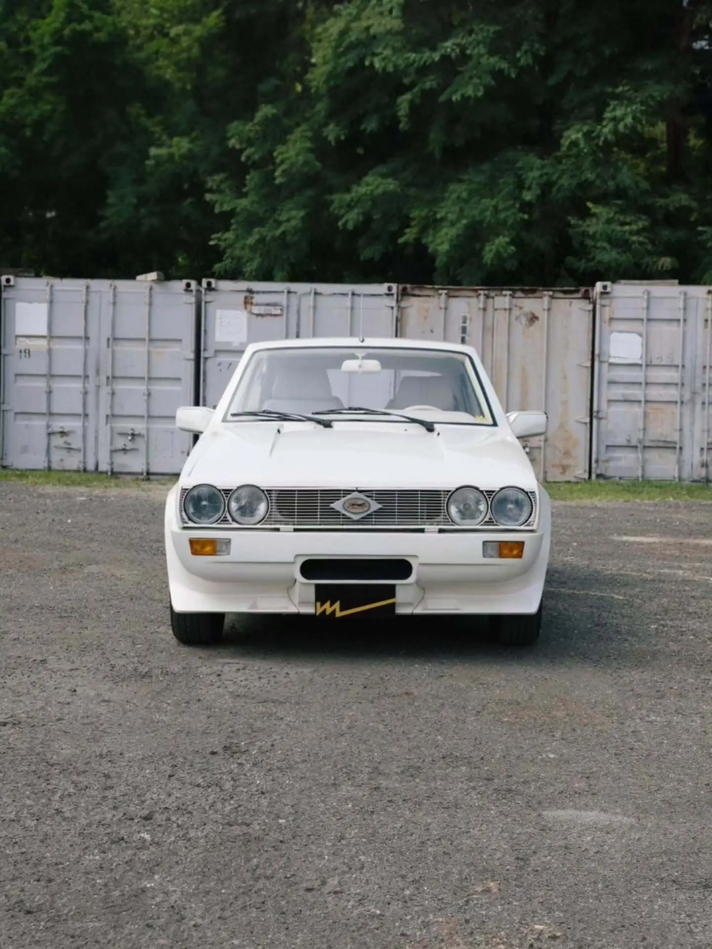 تصویر خودروی کلاسیک و نایاب اسبارو سوپر فایو (Sbarro Super Five) مدل ۱۹۸۵ با بدنه پهن سفید و چراغ‌های جلو چهارگانه در یک منظره کوهستانی.