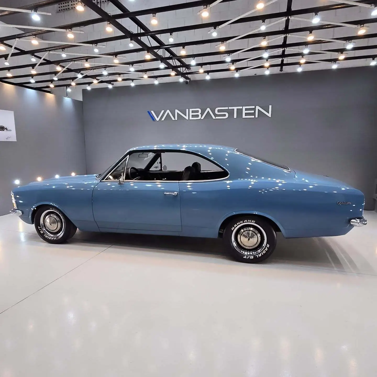 نمای کناری و موتور ارتقایافته شورولت اوپالا ۲۵۰۰ مدل ۱۹۷۶ توربو به رنگ سفید با رینگ‌های کلاسیک و کیت بدنه فابریک در وضعیت نمایشگاهی.