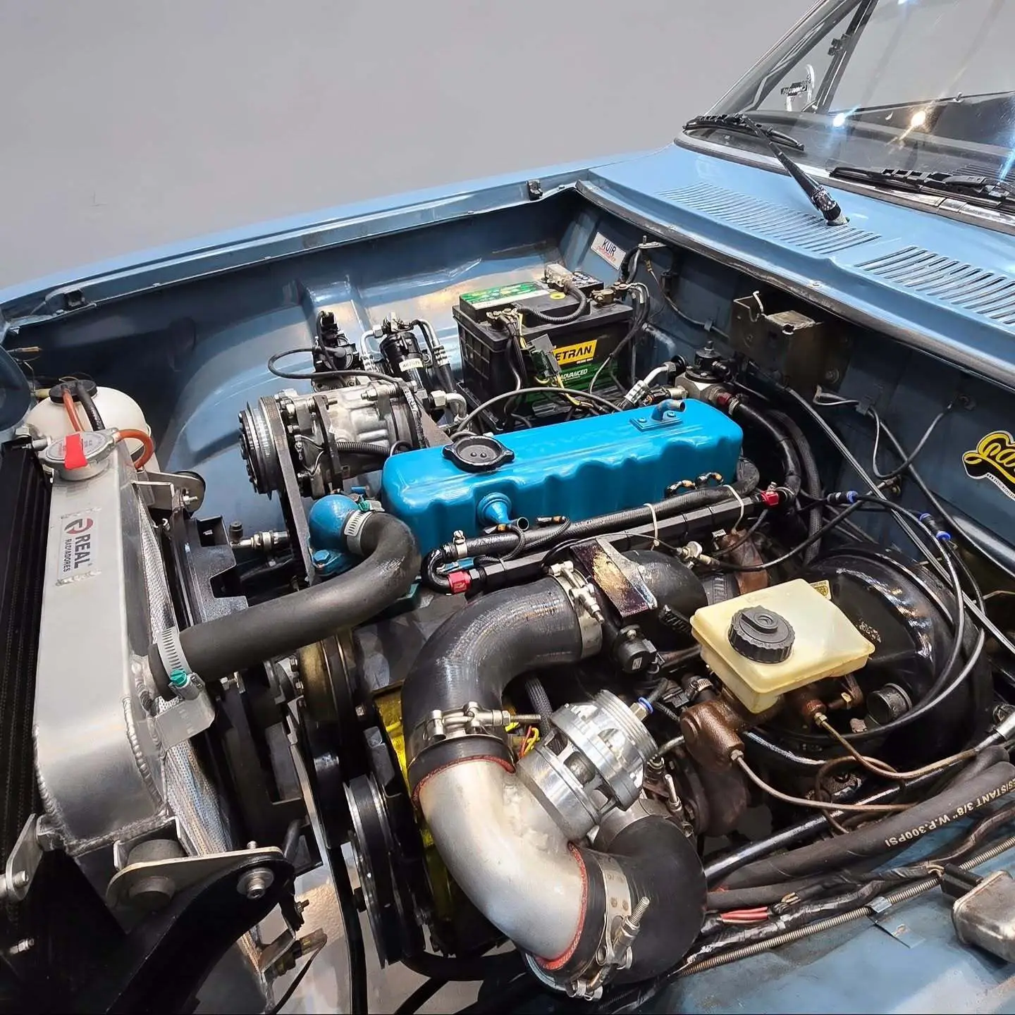 نمای کناری و موتور ارتقایافته شورولت اوپالا ۲۵۰۰ مدل ۱۹۷۶ توربو به رنگ سفید با رینگ‌های کلاسیک و کیت بدنه فابریک در وضعیت نمایشگاهی.