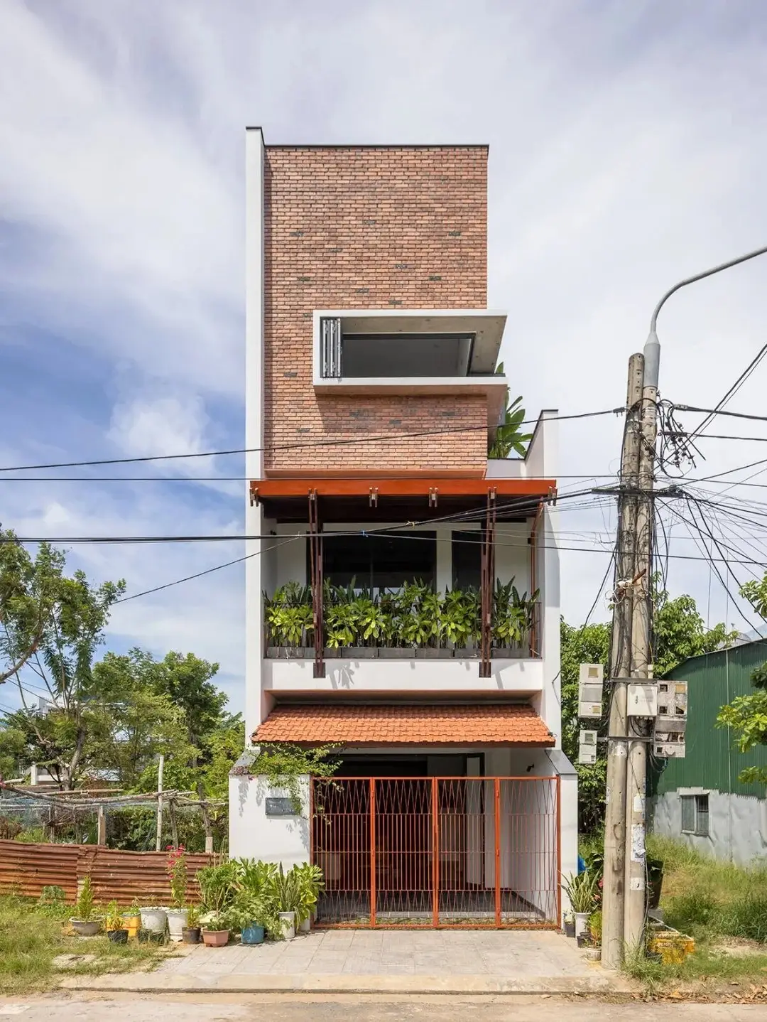 نمای خارجی مدرن خانه برنج قهوه‌ای با ترکیب آجرهای سفالی ترکوتا، بلوک‌های شیشه‌ای رنگی و فضای سبز که نشان‌دهنده معماری پایدار و نورپردازی طبیعی است.