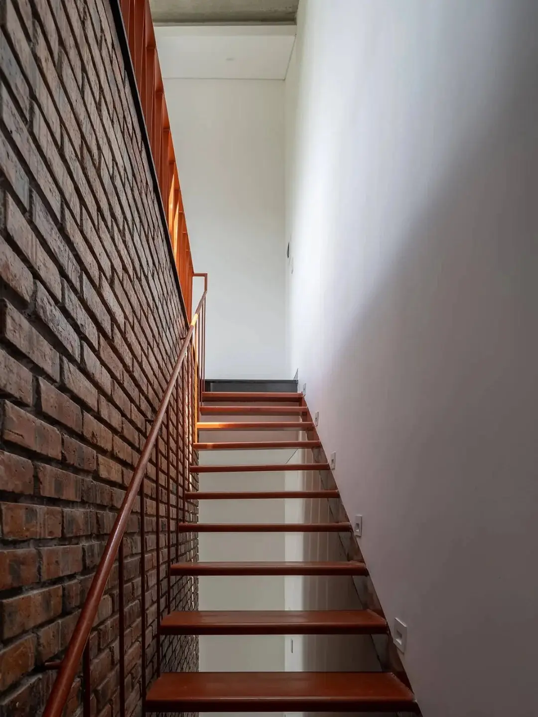 نمای خارجی مدرن خانه برنج قهوه‌ای با ترکیب آجرهای سفالی ترکوتا، بلوک‌های شیشه‌ای رنگی و فضای سبز که نشان‌دهنده معماری پایدار و نورپردازی طبیعی است.