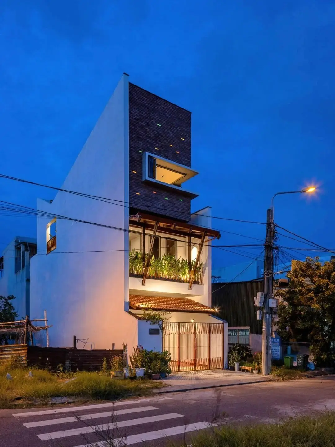 نمای خارجی مدرن خانه برنج قهوه‌ای با ترکیب آجرهای سفالی ترکوتا، بلوک‌های شیشه‌ای رنگی و فضای سبز که نشان‌دهنده معماری پایدار و نورپردازی طبیعی است.