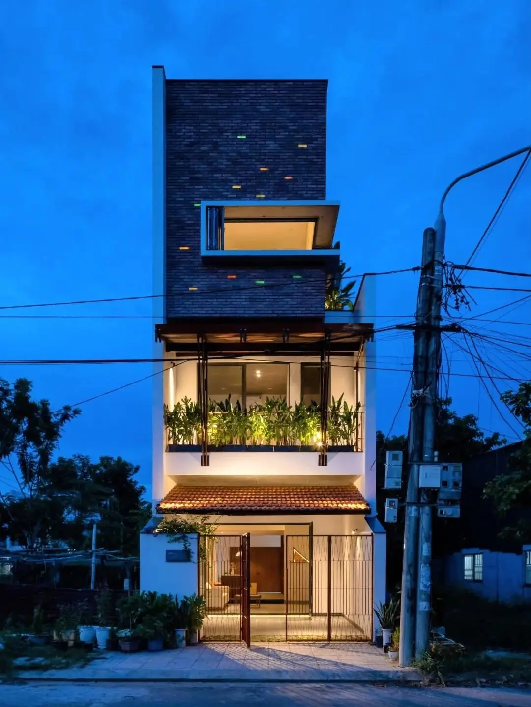 نمای خارجی مدرن خانه برنج قهوه‌ای با ترکیب آجرهای سفالی ترکوتا، بلوک‌های شیشه‌ای رنگی و فضای سبز که نشان‌دهنده معماری پایدار و نورپردازی طبیعی است.