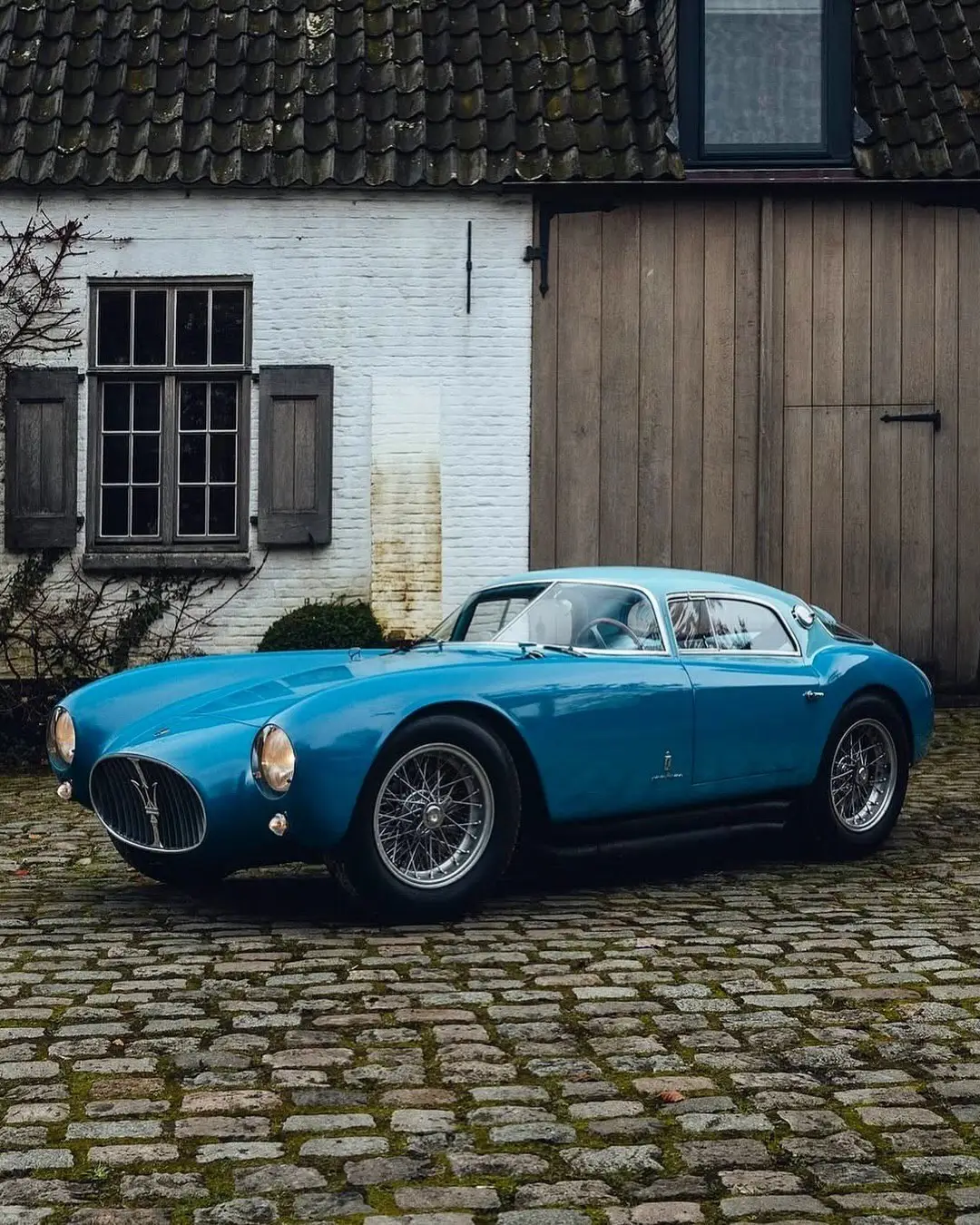 نمای جلویی و جانبی خودروی کلاسیک مازراتی A6GCS فارینا برلینتا به رنگ آبی متالیک با رینگ‌های پره‌ای در مقابل یک بنای سنتی اروپایی.