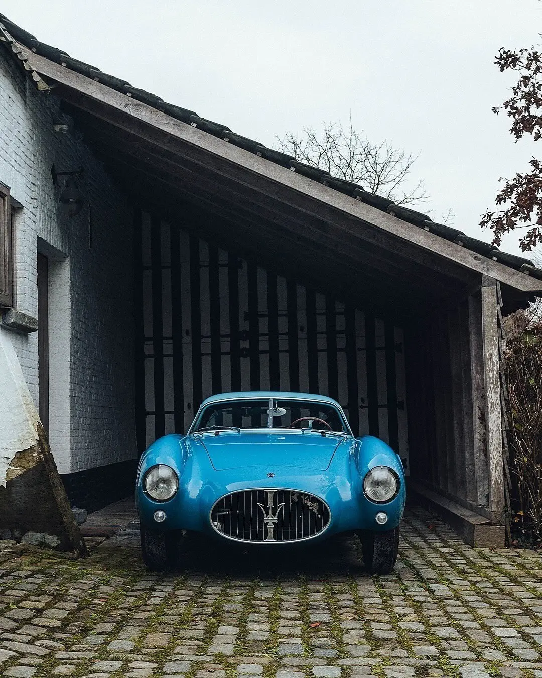 نمای جلویی و جانبی خودروی کلاسیک مازراتی A6GCS فارینا برلینتا به رنگ آبی متالیک با رینگ‌های پره‌ای در مقابل یک بنای سنتی اروپایی.