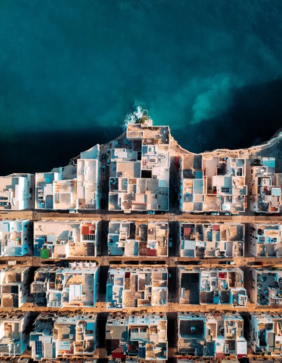 نمای هوایی عمودی از خانه‌های سفید با سقف‌های مسطح در شهر ساحلی پولینیانو آ ماره ایتالیا که در لبه صخره‌های مرتفع کنار دریای آبی قرار گرفته‌اند.