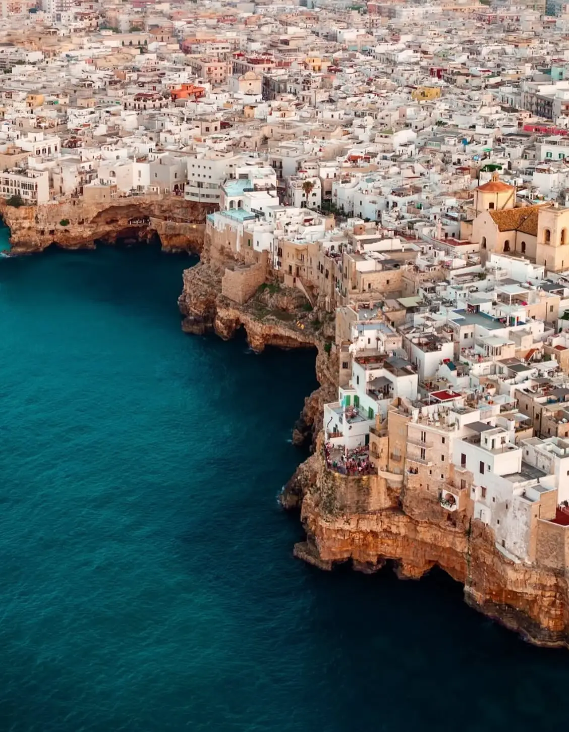 نمای هوایی عمودی از خانه‌های سفید با سقف‌های مسطح در شهر ساحلی پولینیانو آ ماره ایتالیا که در لبه صخره‌های مرتفع کنار دریای آبی قرار گرفته‌اند.