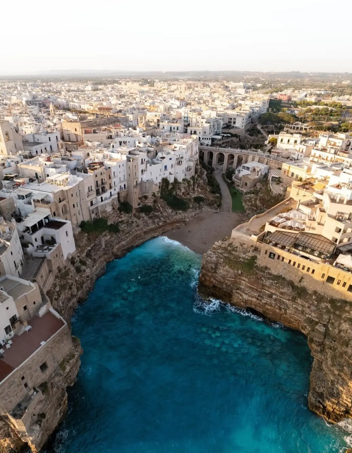 نمای هوایی عمودی از خانه‌های سفید با سقف‌های مسطح در شهر ساحلی پولینیانو آ ماره ایتالیا که در لبه صخره‌های مرتفع کنار دریای آبی قرار گرفته‌اند.