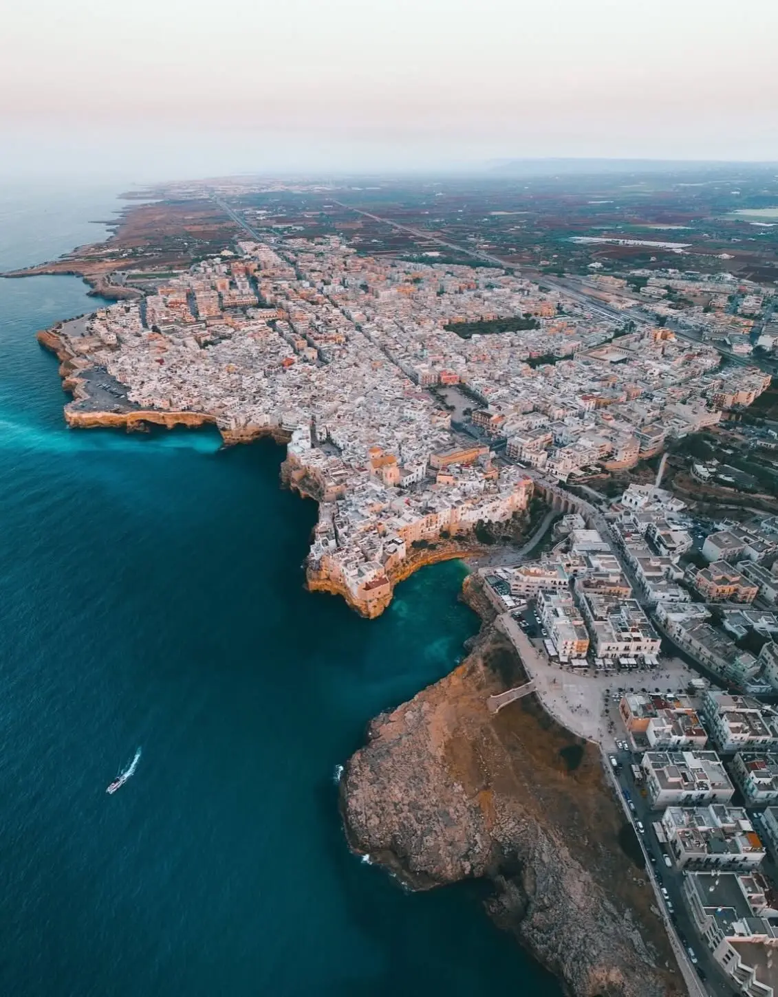 نمای هوایی عمودی از خانه‌های سفید با سقف‌های مسطح در شهر ساحلی پولینیانو آ ماره ایتالیا که در لبه صخره‌های مرتفع کنار دریای آبی قرار گرفته‌اند.