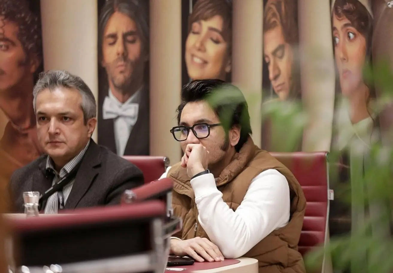 farahang_two_men_talking_on_camera_posters_of_pop_stars_behin_9a030ed4